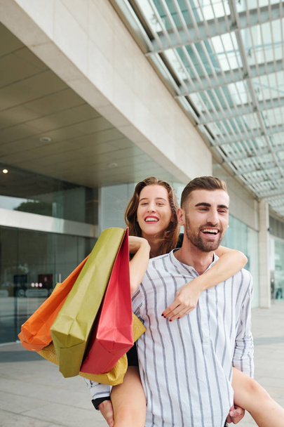 Atractivo joven alegre dando paseo a cuestas a su encantadora novia con muchas bolsas de compras coloridas cuando está de pie en el centro comercial
 - Foto, Imagen