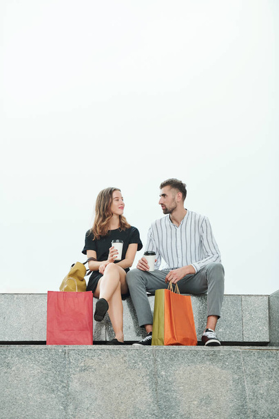Pozytywny młody mężczyzna i kobieta siedzący na granitowych schodach, pijący kawę i omawiający sprzedaż i zakupy - Zdjęcie, obraz