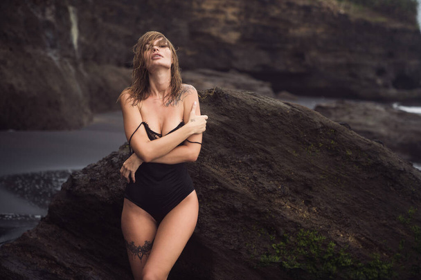 Sexy girl in swimsuit with tattoo on leg rests on black sandy beach on vacation - Zdjęcie, obraz