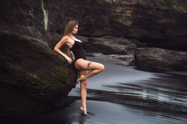 Sexy blond girl in black swimwear rests on black sand beach. - Zdjęcie, obraz