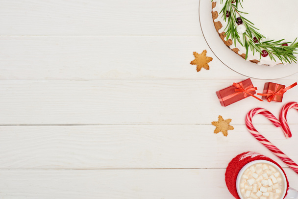 vrchní pohled na vánoční koláč s polevou na bílém dřevěném stole s candy canes, dárky a kakaem - Fotografie, Obrázek