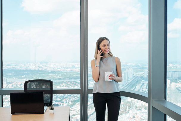 Retrato de una joven mujer de negocios bastante sonriente bebiendo café moderno y llamando por teléfono en la oficina
  - Foto, imagen
