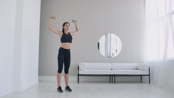 Thuis oefeningen voor een mooi lichaam, een jonge brunette vrouw tilt halters in haar armen in slow motion - Video