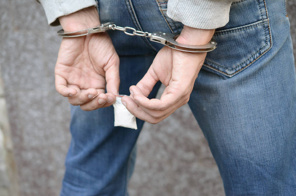 暗い壁の背景に小さなヘロインの薬物パッケージと警察の手錠で逮捕された麻薬ディーラー - 写真・画像