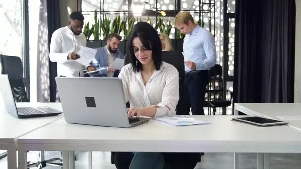 Focused good-looking brunette office worker typing on laptop in the modern office on the background of corporate coworkers - Video