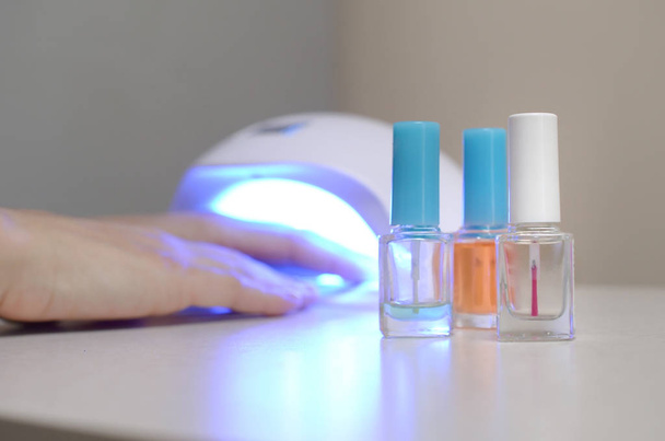 Three colorless transparent nail polish bottles in background of female hands in gel uv led nail white lamp for drying manicure - Photo, Image
