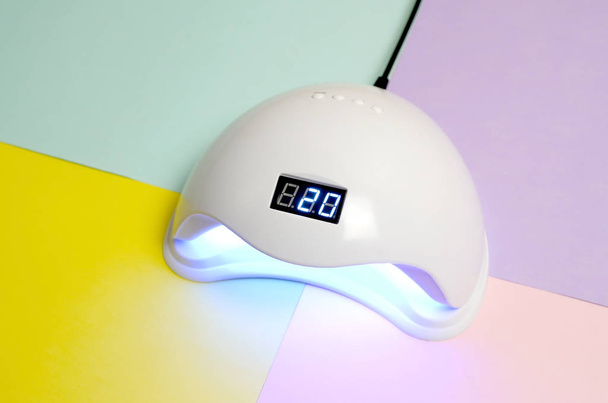 Lâmpada de unha LED UV para processo de cura pelo método de gel encontra-se na mesa multicolorida pastel
 - Foto, Imagem