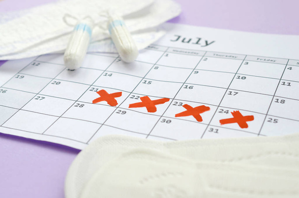 Almohadillas y tampones menstruales en el calendario del período de menstruación con marcas de cruz roja se encuentra en el fondo lila
 - Foto, Imagen
