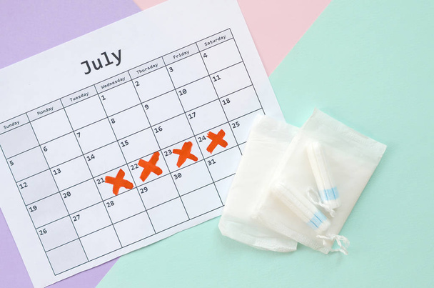 Composición plana con calendario y tampones menstruales y paquetes de almohadillas sobre fondo azul rosado y lila pastel
 - Foto, Imagen