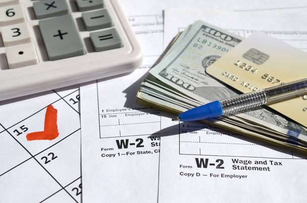 W-2 Заработная плата и налоговая выписка с кредитной картой на долларовых купюрах, калькуляторе и ручке на странице календаря с пометкой 15 апреля
 - Фото, изображение