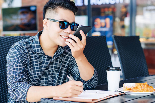 Улыбающийся молодой человек записывает голосовое сообщение во время завтрака и работы за столом кафе
  - Фото, изображение