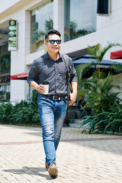 Joven guapo alegre caminando por la calle con una taza de café en la mano
  - Foto, Imagen