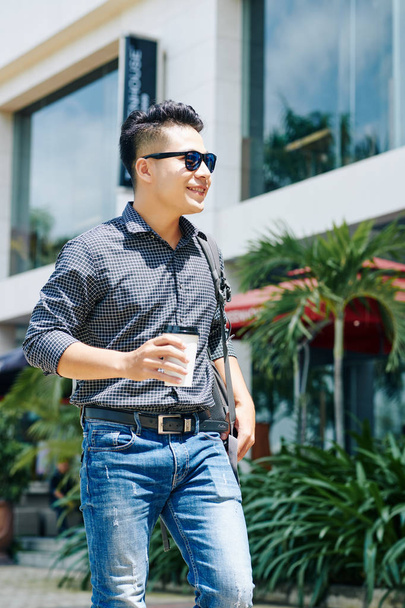 Portrait of smiling young Asian man standing in the street with cup of coffee in hand and enjoying sunny day - Photo, Image