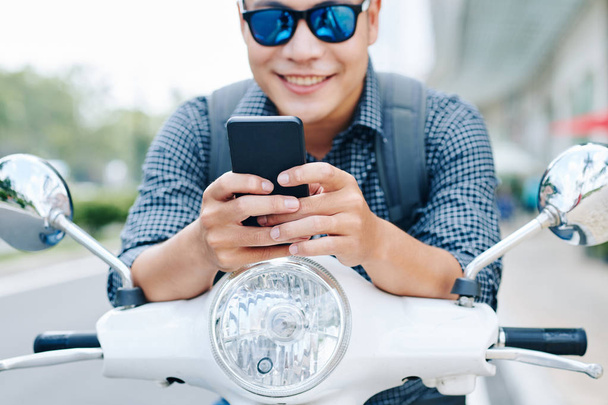 Ευτυχισμένος νεαρός Ασιάτης με γυαλιά ηλίου κάθεται σε σκούτερ και τον έλεγχο των μηνυμάτων κειμένου και ειδοποιήσεις στο τηλέφωνό του, επιλεκτική εστίαση - Φωτογραφία, εικόνα