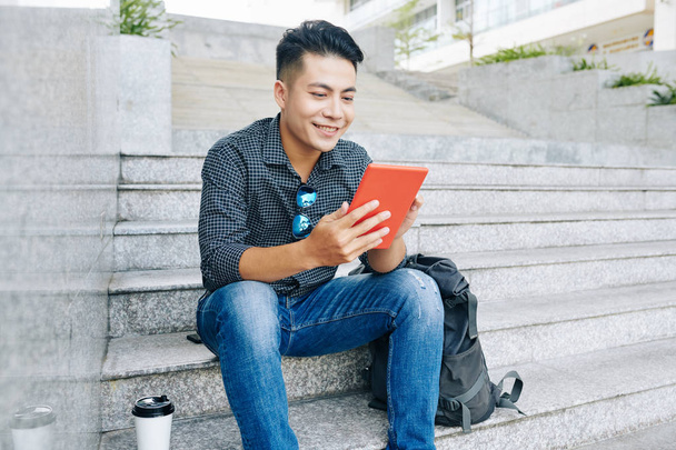 Joven hombre vietnamita positivo sentado en los escalones con una taza de café y artículo de lectura en la computadora de mesa
 - Foto, imagen