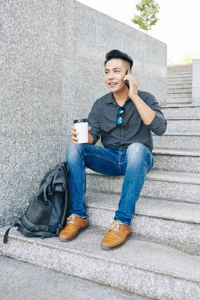 Handsome young Asian man sitting on steps, drinking take away coffee and calling on phone - Photo, Image
