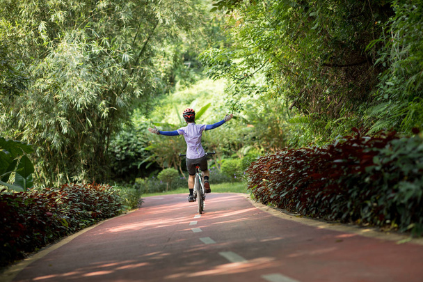 Rear view of female cyclist riding mountain bike on tropical rainforest trail with arms outstretched - Фото, зображення