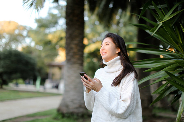 Kiinalainen mukava tyttö käyttää älypuhelinta ja kävely trooppisessa puistossa
. - Valokuva, kuva
