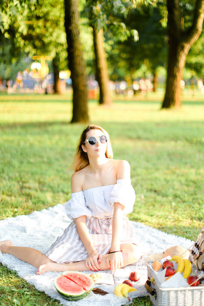 Young european woman in sunglasses having picnic on plaid and sitting in park with fruits. - Photo, image
