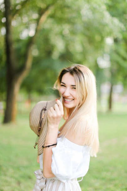Jovem sorrindo mulher de pé no parque e mantendo chapéu
. - Foto, Imagem