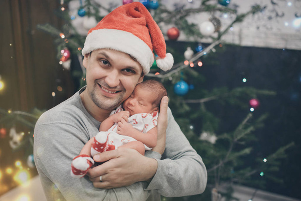 Чоловік у Різдвяному капелюсі тримає новонароджену дитину на задньому плані ялинки в садах. Тато, одягнений як Санта Клаус, засинає. Концепція здорового способу життя, Івф, Крістм - Фото, зображення