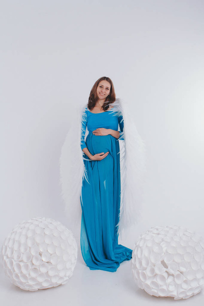 A pregnant girl in a blue dress with angel wings on a white background. An angel-woman have big white balls. The concept of Christmas, mystical creatures, Halloween, new year suit - Photo, Image