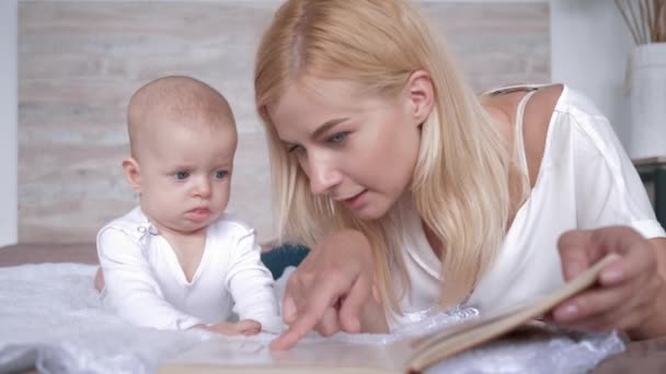 new mother reads a book to her little daughter who listens curiously to her while lying together on bed at home - Footage, Video