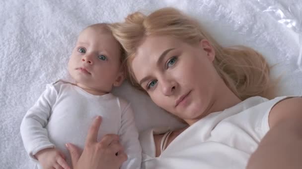 relationship child and mother, young parent with a sweet infant girl lying on white bed and pose on camera close up - Materiał filmowy, wideo