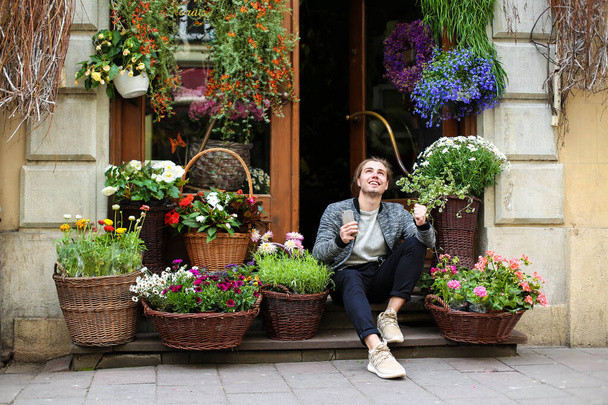 Мальчик держит смартфон и сидит в цветочном магазине рядом с букетами в корзинах
. - Фото, изображение