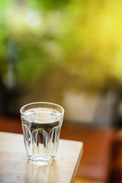 The glass is almost full of water and placed on a light wooden table. Natural green background and a little light for copy space. - Foto, afbeelding