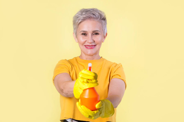 beautiful stylish woman aged, in a yellow T-shirt and rubber gloves on a yellow background with detergent - Foto, imagen