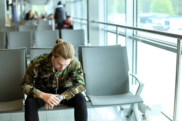 Молодий чоловік сидить у залі очікування в аеропорту, слухає музику з навушниками та дивиться
. - Фото, зображення