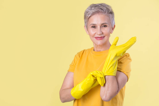 beautiful stylish woman aged, in a yellow T-shirt and rubber gloves on a yellow background - Foto, imagen