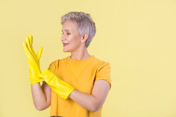 beautiful stylish woman aged, in a yellow T-shirt and rubber gloves on a yellow background - Foto, Imagem