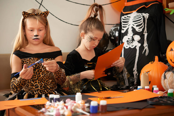 Zwei Mädchen sitzen am Tisch und basteln gemeinsam für den Halloween-Feiertag Figuren aus buntem Papier - Foto, Bild