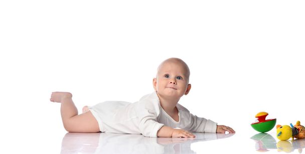 Baby kind baby jongen peuter in wit bodysuit liggend op zijn buik op zoek omhoog kruipen naar kleurrijke speelgoed te krijgen op wit  - Foto, afbeelding