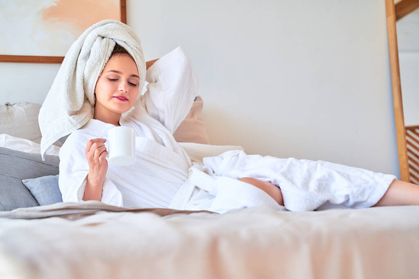 Image of happy woman in white bathrobe with mug of tea in her hands lying on bed. - Foto, immagini