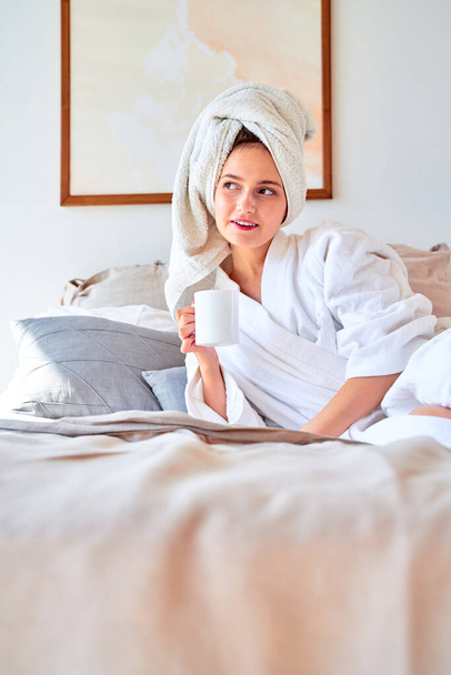 Photo of woman in white bathrobe with mug of tea in her hands lying on bed. - Foto, Imagem