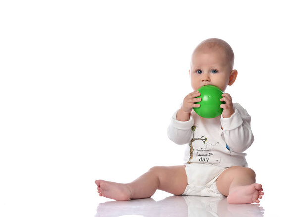 Infant child baby boy  in diaper and white shirt is sitting  with green ball toy in his hands, biting it on white  - Photo, image