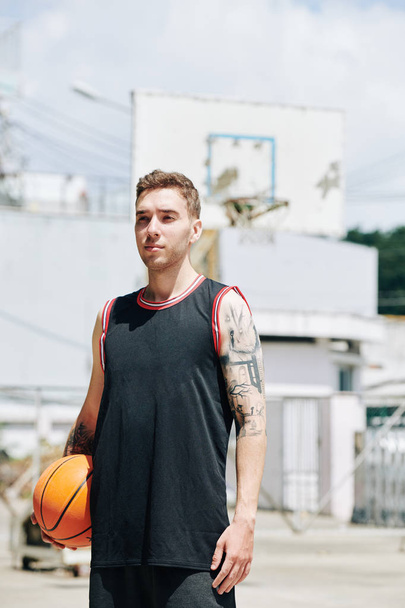 Porträt eines hübschen jungen Basketballspielers, der mit Ball in der Hand auf dem Außenplatz steht - Foto, Bild