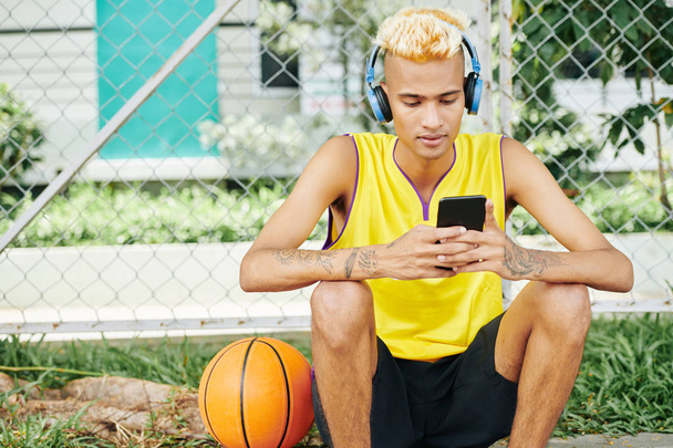 Young handsome man sitting on street basketball court and listening to music in smartphone application via headphones - Photo, Image