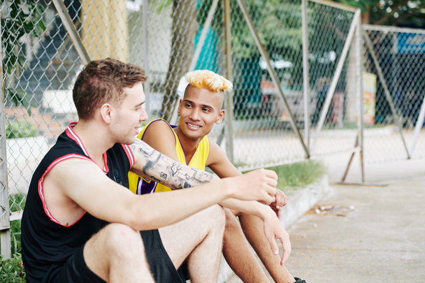 Hymyilevä komea nuori musta mies kuuntelee tarinaa ystävästään, kun he lepäävät pelattuaan urheilupeliä ulkona
 - Valokuva, kuva