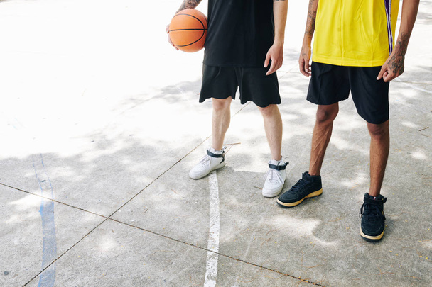 Imagen recortada de jugadores de baloncesto en uniforme negro y amarillo de pie sobre pista de asfalto al aire libre con pelota
 - Foto, Imagen