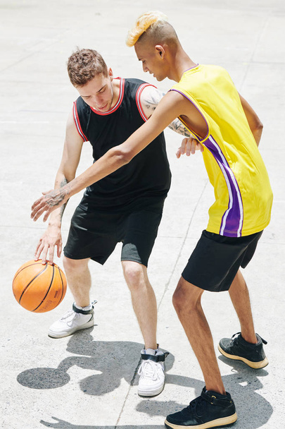 Πολυεθνικοί νέοι με στολή που εξασκούνται στις δεξιότητες μπάσκετ στον αγωνιστικό χώρο την ηλιόλουστη μέρα - Φωτογραφία, εικόνα