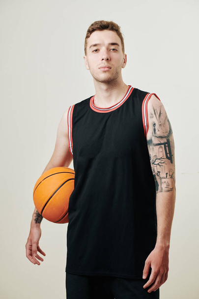 Sebevědomý seriózní basketbalista v černé uniformě drží míč a dívá se na kameru, izolované na bílém - Fotografie, Obrázek