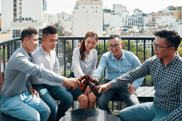 Aziatische collega 's uit het bedrijfsleven in casual kleding zitten en toasten met flessen bier tijdens hun bijeenkomst in outdoor cafe - Foto, afbeelding
