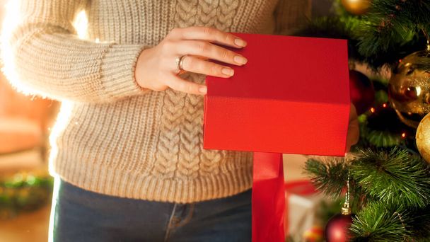 Closeup image of woman standing next to Christmas tree and opening red gift box - 写真・画像