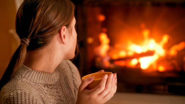 Portrait of woman in woolen sweater holding cup of hot tea and looking at burning firepalce - Photo, Image