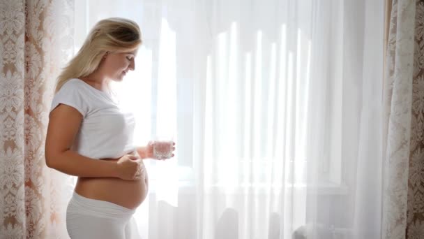 Şanslı hamile kadın cam bardağı temiz su tutuyor ve pencerenin yanında kocaman çıplak göbeği ovuyor. - Video, Çekim