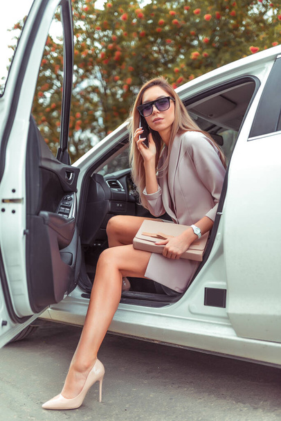 woman gets out car summer autumn city. Taxi business class. In hand mobile phone, car sharing rental, pink suit high-heeled shoes. Tanned slender girl with long hair. Business important meeting. - Fotografie, Obrázek